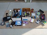 Wide overview of two boxes and assortment of office supplies including calculators, tape dispensers, stamps, pens, and organizers arranged outdoors on mat.