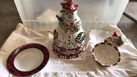 Photo showing the Tracy Porter snowman cookie jar with a blue bird on its arm and holly details, next to a ceramic bowl with red and green holly rim and a china plate with tartan plaid rim, placed on light fabric background.