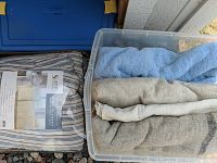 Photo showing 5 blankets in a plastic tub and on the ground, including wool and cotton blankets in various colors, one packaged.