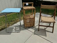 Three items outdoors on a paved surface: aqua patterned folding table, wicker shelving unit, wicker folding chair.