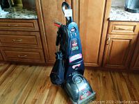 Full view of the Bissell ProHeat Turbo 2X carpet shampooer standing upright on a wooden floor near kitchen cabinetry. Black mesh bag is attached to the unit.