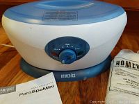 Homedics ParaSpaMini electric paraffin bath unit with control knob and indicator lights. Instruction booklet and part of wax package shown nearby.