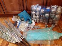 Top-down view showing clear plastic packages of silver and blue ornaments, rolls of light blue fabric with silver snowflakes, silver glitter flower stems, large blue glitter snowflakes, and silver pinecone picks all arranged on wood floor.