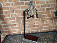 Full side-angle view of the vintage metal wine bottle opener with ornate grapevine design and metal stand on a table against a brick wall.