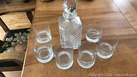 Full view of the crystal decanter with faceted stopper and six matching cocktail rocks glasses arranged around it on a wooden table.