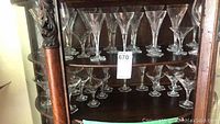 Wide view of multiple clear crystal wine, champagne, and cocktail glasses arranged on two shelves in a wooden cabinet. Glasses have hexagonal bases.