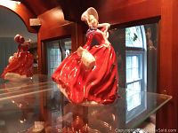 Side view of Royal Doulton figurine in red dress with bonnet and basket, displayed on glass shelf.