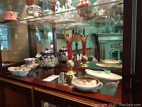 Wide view showing multiple fine china pieces including lidded bowls, dishes, teacup and saucer, and the red and gold gilded vase inside glass display cabinet.