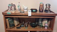 Wide angle photo showing the top of wooden cabinet with the variety of items arranged including the lighted cottage, figurines, mortar and pestle, and decorative boxes