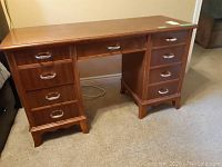 Front view of a medium brown wood desk with eight drawers and metal pull handles. Surface marks visible on top.