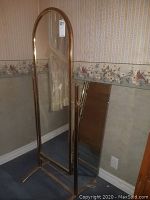Full view of the free-standing brass framed floor mirror with arched top, showing side supports and base on carpeted floor near wallpapered wall.