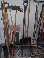 Photo showing multiple shovels, garden rake with orange handle, and push brooms with wooden handles.