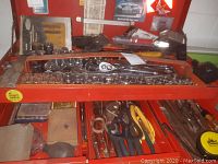 Top view of open red metal toolbox showing rows of chrome sockets, ratchet handles, screwdrivers, pliers, measuring tape, and other tools inside.
