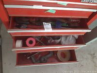 Partial view of three red toolbox drawers with various hand tools like pliers, tape rolls, and measuring tools inside.