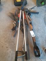 Overhead view of assorted garden tools laid on garage floor, showing multiple rakes, clippers, and weed pullers with wooden and metal handles.