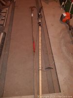 Two long-handled tree pruning tools laid out side by side on a carpet, one is a limb trimmer with a saw blade and the other a limb lopper with a hooked cutting blade.