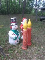Wide shot showing all four outdoor Christmas decorations on grass near trees: snowman, Santa holding HOHOHO sign, and two NOEL candles.