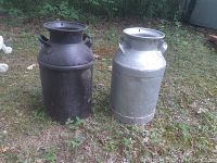 Two vintage milk cans positioned outdoors on grass. One black, one silver with visible hole on silver can bottom edge.