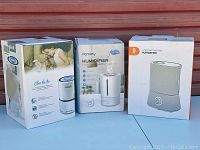 Three humidifiers in front of metal door: Levoit, Homasy and Ultrasonic Color Mist with packaging
