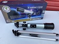 Photo showing the telescope with optical tube, diagonal prism and aluminum tripod stand alongside the product box labeled F36050.