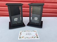 Pair of black rectangular outdoor lanterns with glass panels and small white candles inside, placed on flat surface with decorative 'Emclora thank you' card.
