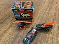 Box of 1985 Topps Batman Deluxe Reissue Edition trading cards with 143 cards visible on box, beside three Batman vehicle toys: Batmobile, Batboat and Batcopter
