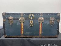 Front view of vintage trunk showing metal latch locks, leather straps with red detailing, metal reinforcements on corners and edges.