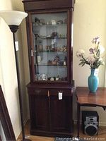Full view of wooden curio cabinet with glass upper door showing display items inside, situated next to a floor lamp and small table with artificial flowers.