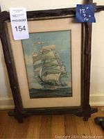 Full view of framed sailing ship art resting against wall on wooden floor. The frame has a rustic textured carved leaf design.