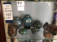 Displays seven decorative boxes and jars on a glass shelf including blue and green cloisonné pieces and wooden boxes.