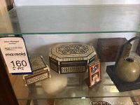 Glass shelf displaying five small wooden decorative boxes with intricate inlaid geometric patterns and one small round vase with handle.