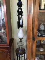 Full view showing the black macramé hanging, the vintage glass oil lamp resting on a small round metal table with wooden top, positioned between wooden cabinets.