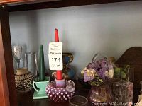 Photo shows purple hobnail glass candle holder with red candle, green ceramic candle holder, and clear glass basket with faux flowers on wooden shelf along with additional glass and ceramic decorative objects.