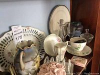 Photo showing ceramic plates, teacups, porcelain pitchers, green glass stopper bottle, and decorative ceramic flower bouquet arranged together.