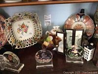 Shelf display of two large ornate decorative plates, decorative glass pieces with bird and floral themes, miniature folding screen with Asian landscape paintings, lidded ceramic dish, and small bottles. Includes the oriental vase partially shown at the right edge.