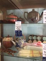 Close-up of glass curio cabinet shelf with assorted teapots, glass and ceramics, plus linens.