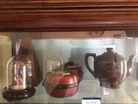 View of musketeer figurine under glass dome, metronome, and ceramic teapot on shelf