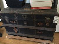 Front view of a large old black trunk showing metal bands, rivets, latches, and leather straps mounted on a wooden frame with wheels.