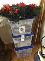 Three clear plastic storage bins stacked, showing they contain various Christmas decorations with blue lids