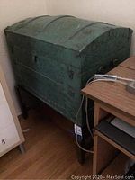 Front side of green metal dome-top trunk on wooden stand showing embossing and latch.