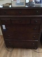 Front view of a dark wooden chest with five drawers and round metal pulls, showing wood grain and surface wear.