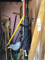 Photo showing various gardening and cleaning tools leaning against a pink wall and wooden door interior, including shovels, rakes, mop, broom, and umbrella.
