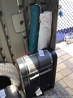 Photo showing the black ceramic heater and two outdoor umbrellas in carrying bags, placed against a wall and near a tire.