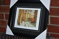 Professional image showing the framed artwork with corner protectors on the black frame, depicting an urban street scene in autumn or early winter with snow on the ground.