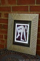 Framed and matted print displayed outdoors against brick background, showing entire frame and matting with textured design.