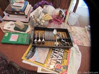 Overview of the entire lot laid out on a wooden dining table with floral tablecloth, showing the cutlery set in its container, stacks of books and maps, glass items, candle, and other small trinkets.