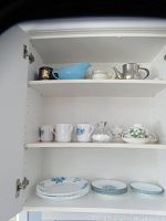 Photo showing shelves inside cupboard with assorted dishes including cups, a blue pitcher, teapot, and covered dish.
