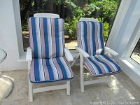 Front view of two white folding plastic lawn chairs with striped cushions on tiled floor by window.