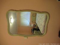 Front view of French Provincial style mirror hung on a peach-colored wall reflecting bedroom furniture.