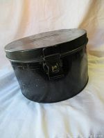 Side angle photo showing the black metal oval hat box with hinged lid and metal latch closure.